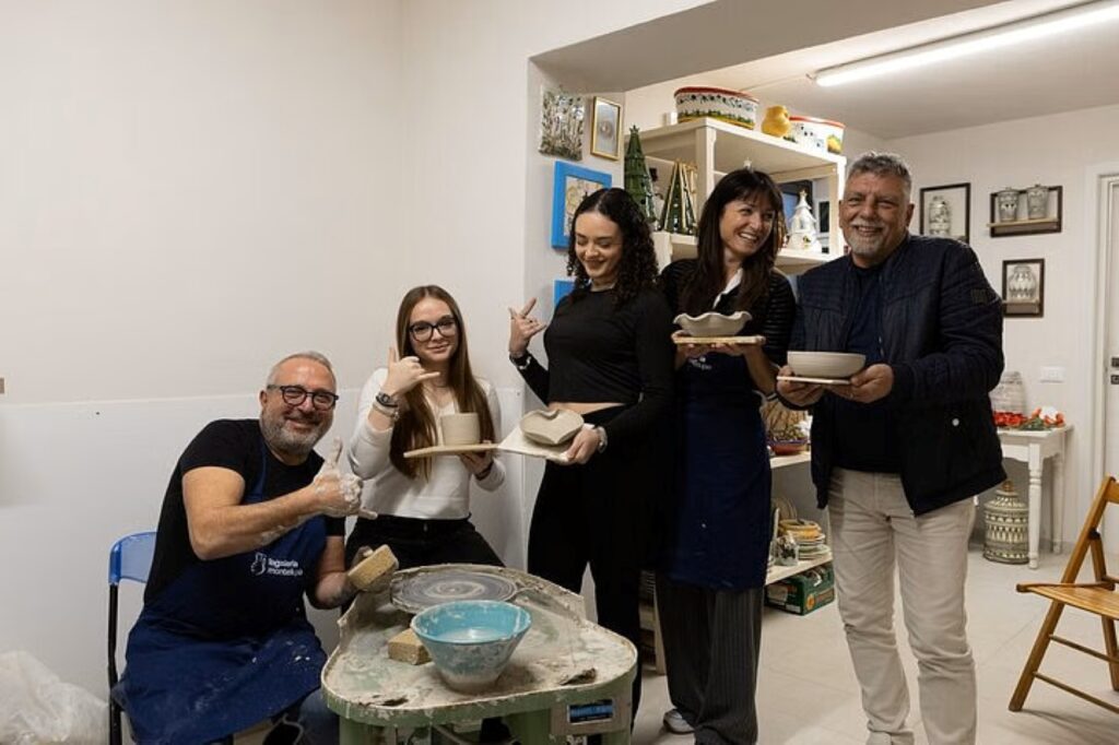 A group of students holding their pottery creations during a Tuscan ceramics masterclass, with a pottery wheel and shelves in the background.