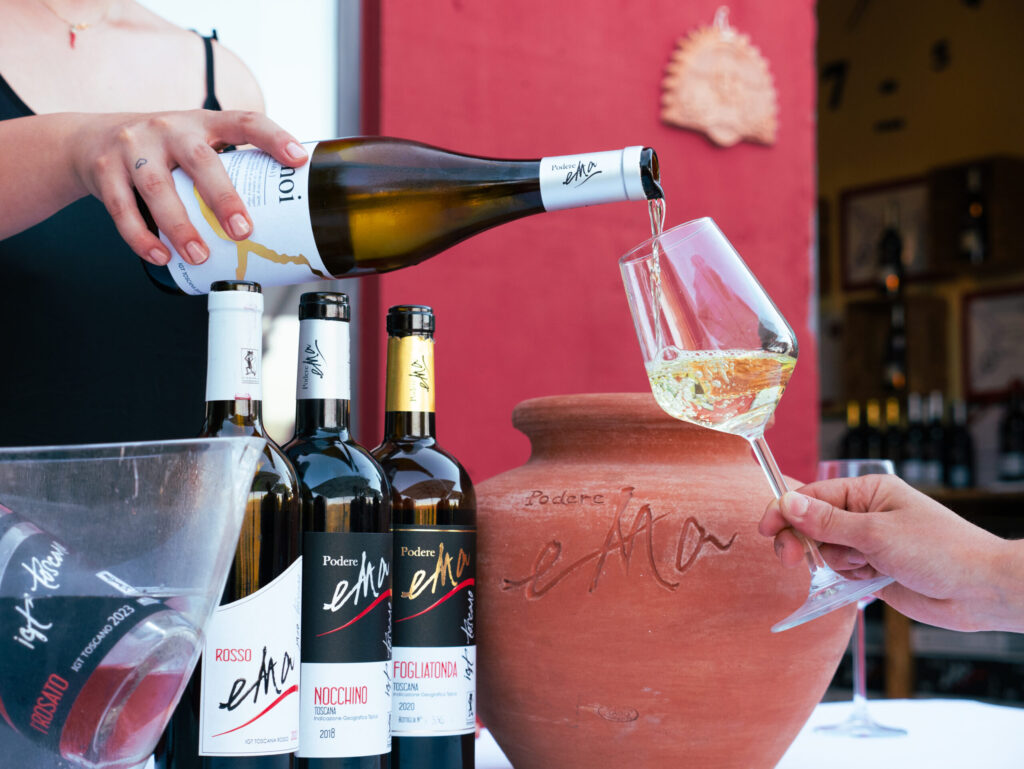 White wine being poured into a glass beside bottles and a terracotta amphora at a Tuscan winery