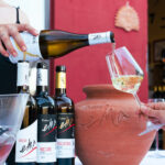 White wine being poured into a glass beside bottles and a terracotta amphora at a Tuscan winery