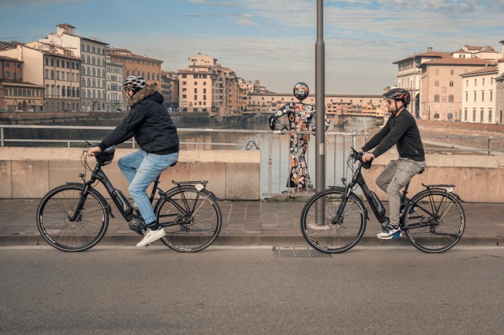 Two riders on an e-bike tour in Florence cycling along the Arno River with Ponte Vecchio in the background