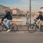Two riders on an e-bike tour in Florence cycling along the Arno River with Ponte Vecchio in the background