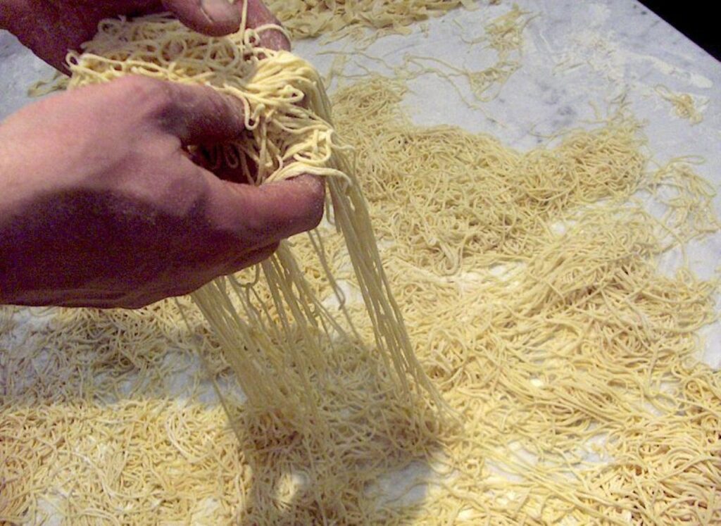 Hand lifting fresh handmade vermicelli pasta strands on a marble work surface.