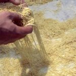 Hand lifting fresh handmade vermicelli pasta strands on a marble work surface.