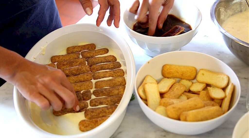 Hands assembling ladyfingers and cream while preparing tiramisu in a Tuscan farmhouse cooking class.