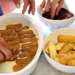Hands assembling ladyfingers and cream while preparing tiramisu in a Tuscan farmhouse cooking class.